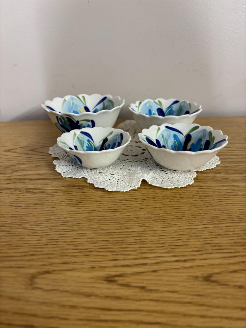 Puede incluir: Cuatro cuencos de cer&aacute;mica blanca con bordes festoneados, cada uno adornado con un dise&ntilde;o floral azul. Los cuencos est&aacute;n dispuestos sobre un tapete de encaje blanco, sobre una superficie de madera clara. El patr&oacute;n floral presenta tonos de azul y verde.