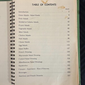 May include: A vintage cookbook with a green cover and a spiral binding. The table of contents lists various salad recipes, including green salads, fruit salads, molded salads, frozen salads, vegetable salads, meat salads, chicken salads, fish salads, cheese salads, egg salads, salad buffet, French dressing, mayonnaise salad dressing, cooked salad dressing, miscellaneous salad dressings, sandwiches, canapes, appetizers, hors d'oeuvres, beverages, and American and French desserts.