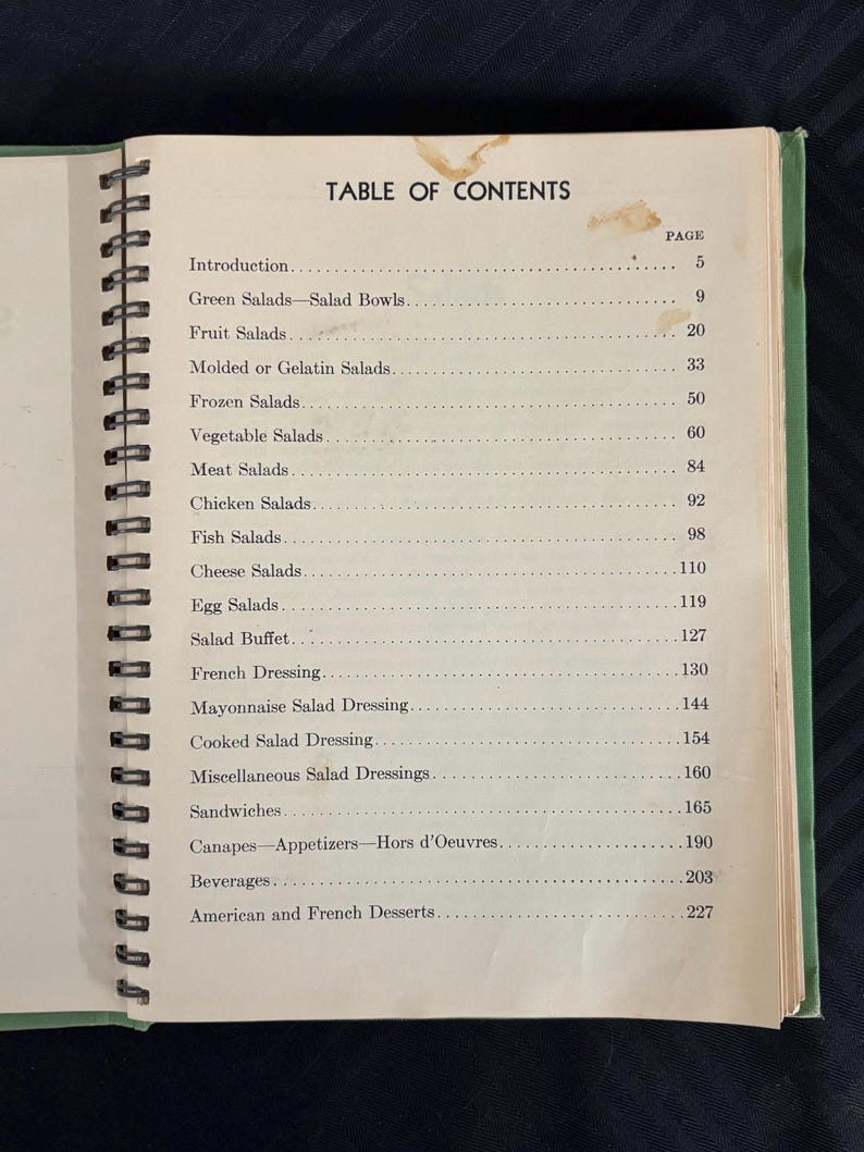 May include: A vintage cookbook with a green cover and a spiral binding. The table of contents lists various salad recipes, including green salads, fruit salads, molded salads, frozen salads, vegetable salads, meat salads, chicken salads, fish salads, cheese salads, egg salads, salad buffet, French dressing, mayonnaise salad dressing, cooked salad dressing, miscellaneous salad dressings, sandwiches, canapes, appetizers, hors d'oeuvres, beverages, and American and French desserts.