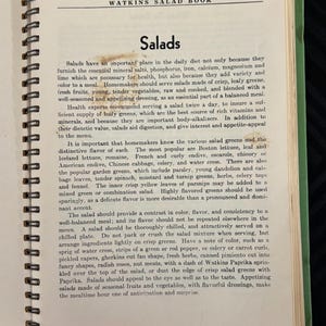 May include: A vintage cookbook titled "Watkins Salad Book" with a green cover and a spiral binding. The page is open to a chapter titled "Salads" and discusses the importance of salads in a balanced diet, including the essential minerals and vitamins they provide. The text also highlights the importance of variety and color in salads, recommending the use of leafy greens, fresh fruits, and vegetables.