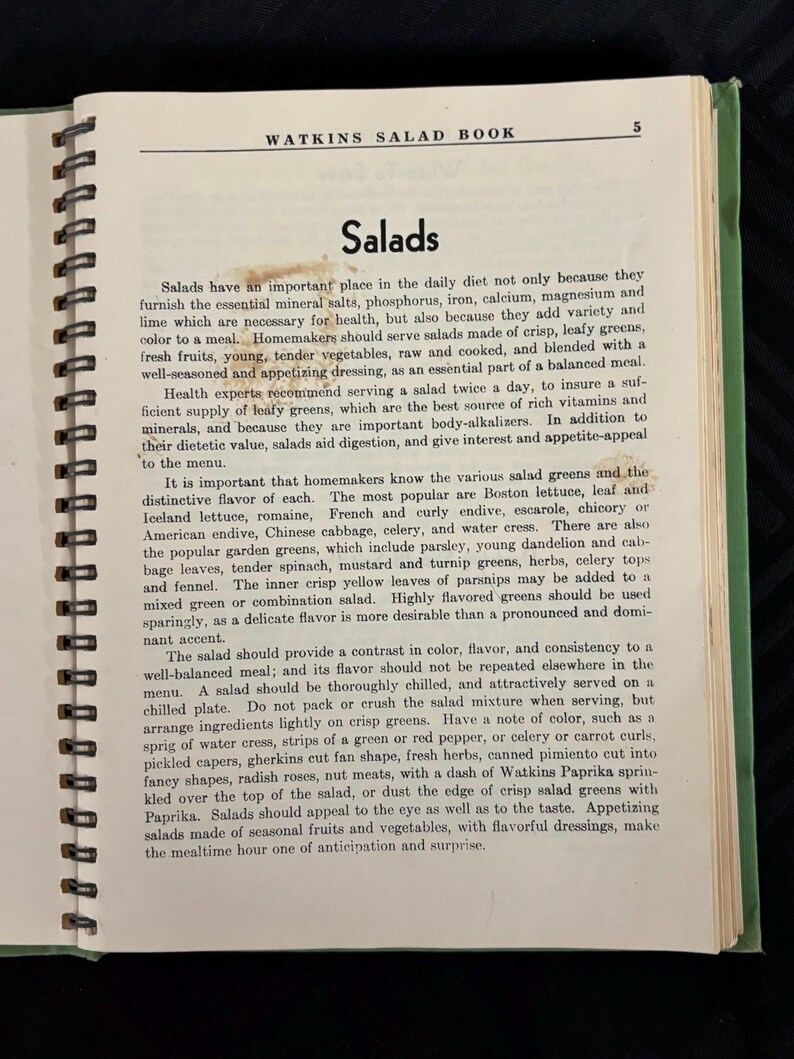 May include: A vintage cookbook titled "Watkins Salad Book" with a green cover and a spiral binding. The page is open to a chapter titled "Salads" and discusses the importance of salads in a balanced diet, including the essential minerals and vitamins they provide. The text also highlights the importance of variety and color in salads, recommending the use of leafy greens, fresh fruits, and vegetables.