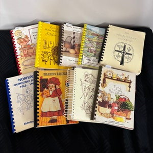 May include: A collection of nine spiral bound cookbooks with various covers. The covers feature illustrations of kitchens, houses, food, and religious symbols. The titles include "Thirty Years of Good Cooking", "Kitchen", "The Presbyterian Cookbook", "Sharing Recipes", "Food for my Household", and "Norfleet Elementary School 1963-1981".