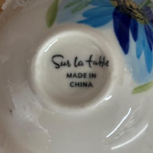 Puede incluir: Platillo de cer&aacute;mica blanca con un dise&ntilde;o floral azul y hojas verdes. Las palabras "Sur la table" y "MADE IN CHINA" est&aacute;n impresas en la parte inferior. El platillo tiene un borde ligeramente curvado.