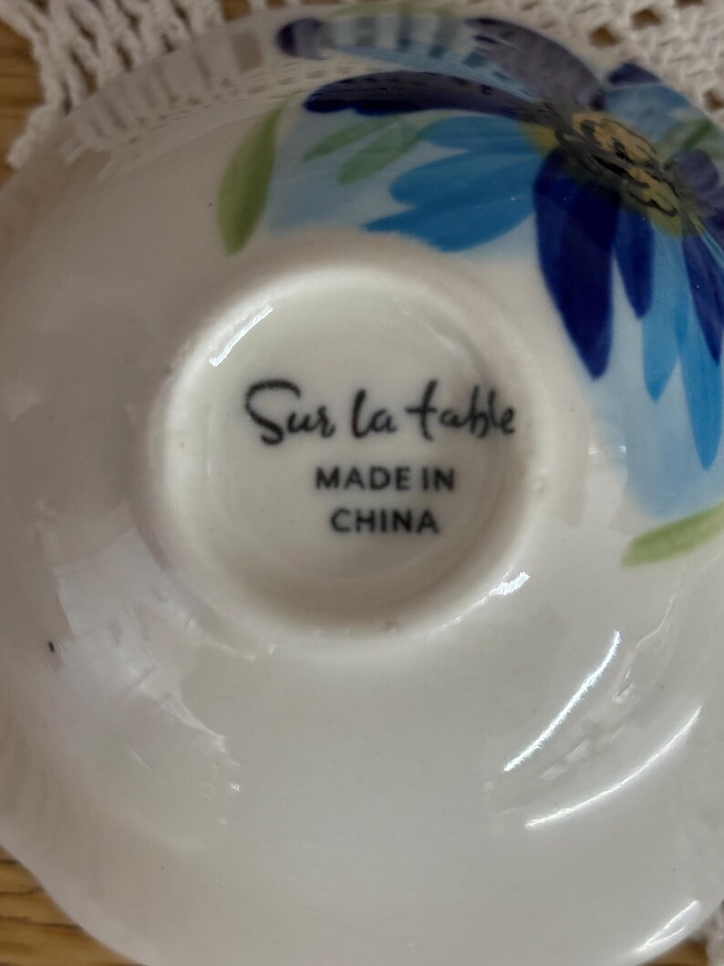 Puede incluir: Platillo de cer&aacute;mica blanca con un dise&ntilde;o floral azul y hojas verdes. Las palabras "Sur la table" y "MADE IN CHINA" est&aacute;n impresas en la parte inferior. El platillo tiene un borde ligeramente curvado.