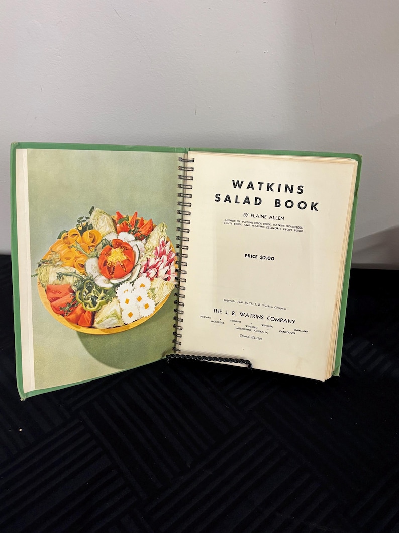 May include: A green and white cookbook titled "Watkins Salad Book" by Elaine Allen. The book is open to a page with a colorful salad in a bowl. The price is $2.00.