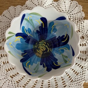 Puede incluir: Un cuenco de cer&aacute;mica blanco con borde festoneado, pintado a mano con un gran dise&ntilde;o de flor azul. La flor tiene diferentes tonos de azul, amarillo y verde. El cuenco est&aacute; sobre un tapete de ganchillo blanco.