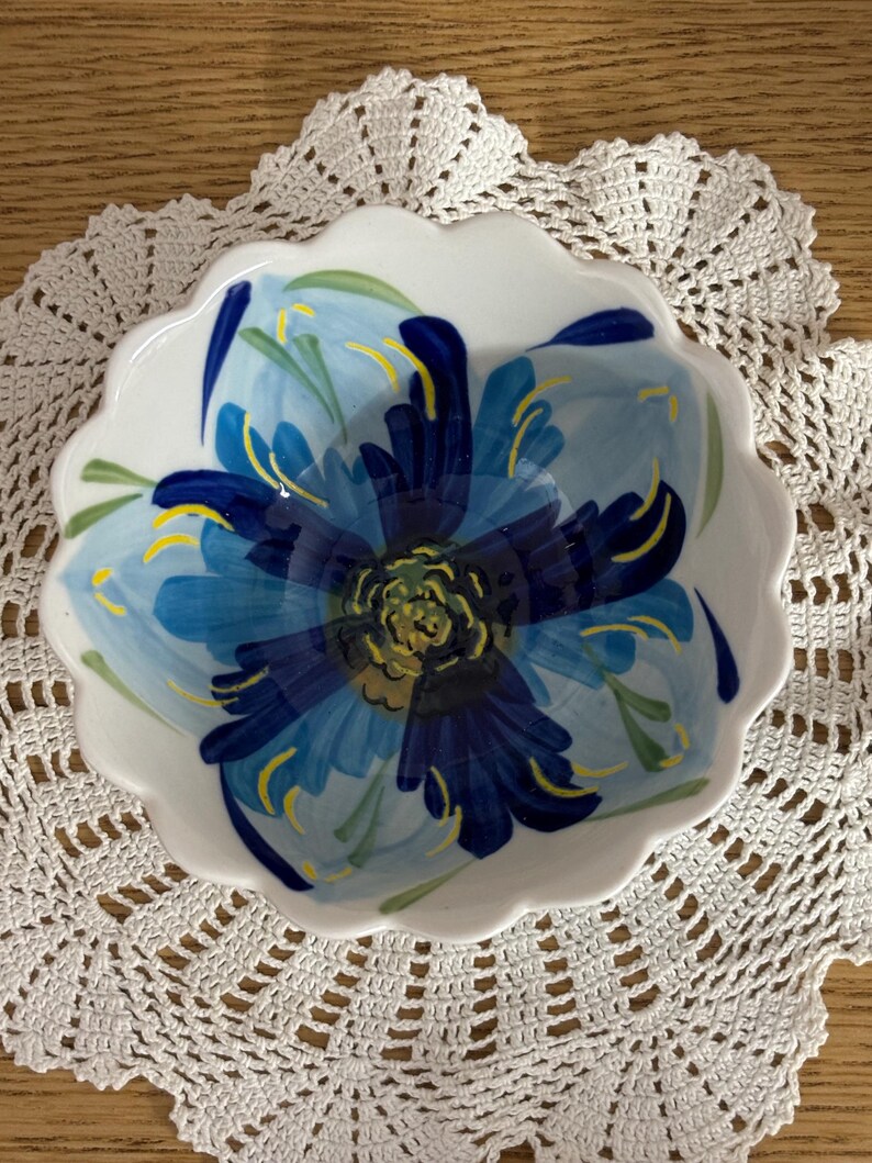 Puede incluir: Un cuenco de cer&aacute;mica blanco con borde festoneado, pintado a mano con un gran dise&ntilde;o de flor azul. La flor tiene diferentes tonos de azul, amarillo y verde. El cuenco est&aacute; sobre un tapete de ganchillo blanco.