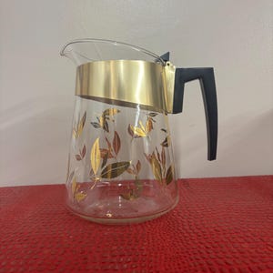 May include: A clear glass pitcher featuring a gold-colored metal band around the top rim and a black plastic handle. The exterior of the glass body is decorated with a repeating pattern of gold and brown leaf illustrations scattered across the surface.