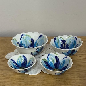 Puede incluir: Cuatro cuencos de cer&aacute;mica blanca con borde festoneado, cada uno adornado con un dise&ntilde;o floral azul. Las flores tienen p&eacute;talos azul oscuro y azul claro con acentos verdes. Los cuencos est&aacute;n dispuestos sobre un tapete de encaje blanco.