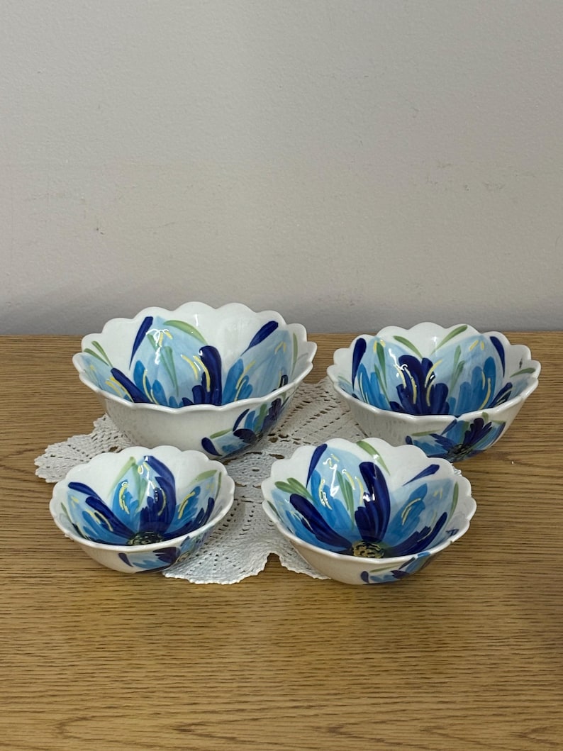 Puede incluir: Cuatro cuencos de cer&aacute;mica blanca con borde festoneado, cada uno adornado con un dise&ntilde;o floral azul. Las flores tienen p&eacute;talos azul oscuro y azul claro con acentos verdes. Los cuencos est&aacute;n dispuestos sobre un tapete de encaje blanco.