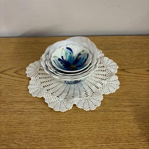 Puede incluir: Un conjunto de cuatro cuencos de cer&aacute;mica blanca con un dise&ntilde;o floral azul, colocados sobre un tapete de ganchillo blanco. Los cuencos tienen un borde festoneado y tama&ntilde;os variables. El dise&ntilde;o floral presenta tonos de azul y verde.