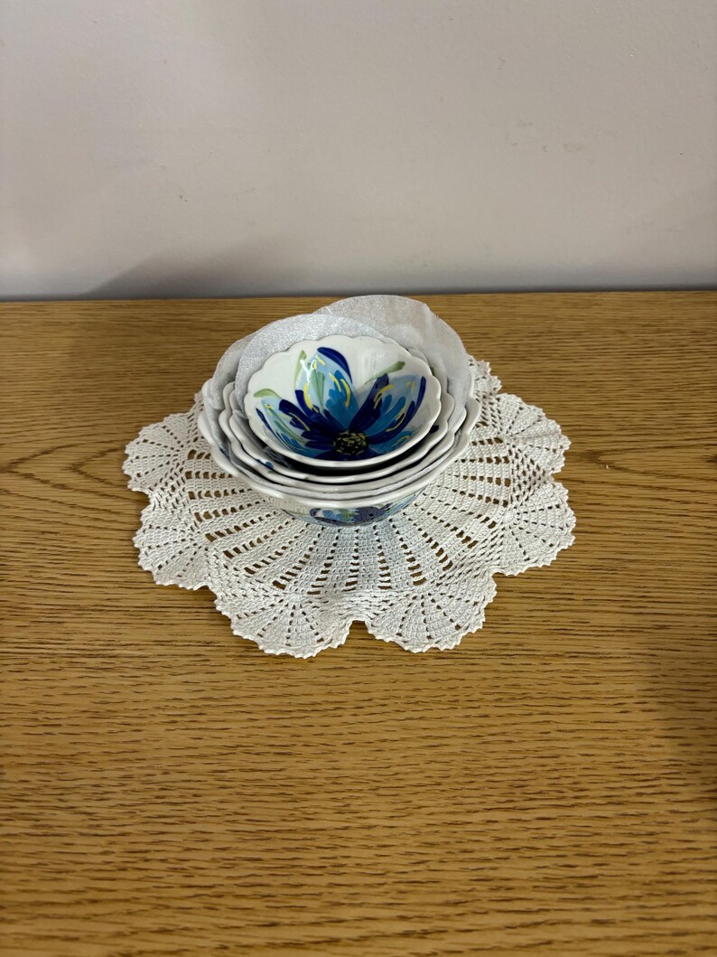 Puede incluir: Un conjunto de cuatro cuencos de cer&aacute;mica blanca con un dise&ntilde;o floral azul, colocados sobre un tapete de ganchillo blanco. Los cuencos tienen un borde festoneado y tama&ntilde;os variables. El dise&ntilde;o floral presenta tonos de azul y verde.