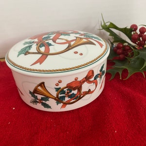 May include: White porcelain trinket box with a Christmas theme. The lid features a gold and red ribbon with a pair of gold horns and holly leaves. The sides of the box feature a similar design with a gold horn, red ribbon, and holly leaves.