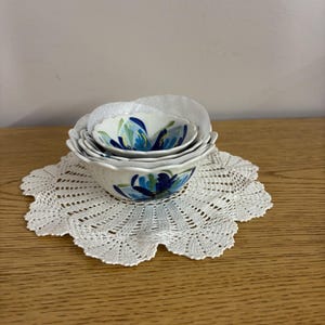 Puede incluir: Un juego de cuatro cuencos de cer&aacute;mica blanca con un dise&ntilde;o floral azul, anidados sobre un tapete de ganchillo blanco. Los cuencos var&iacute;an en tama&ntilde;o, con el cuenco m&aacute;s grande en la parte inferior. El tapete tiene un borde festoneado.