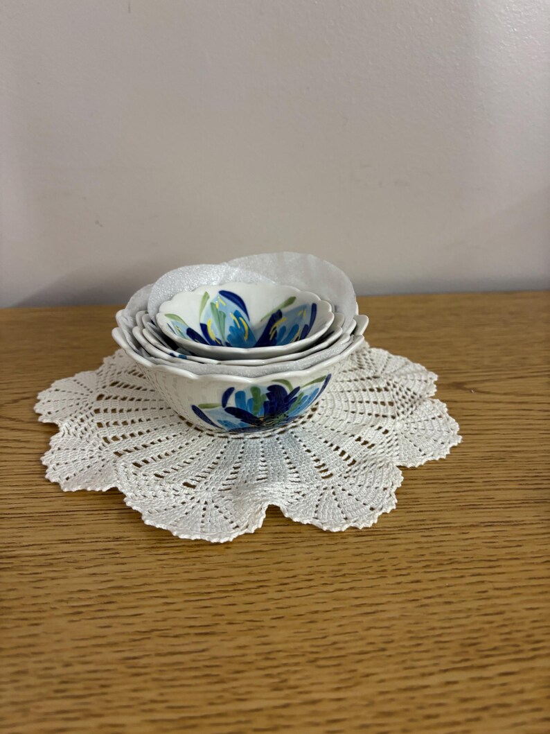 Puede incluir: Un juego de cuatro cuencos de cer&aacute;mica blanca con un dise&ntilde;o floral azul, anidados sobre un tapete de ganchillo blanco. Los cuencos var&iacute;an en tama&ntilde;o, con el cuenco m&aacute;s grande en la parte inferior. El tapete tiene un borde festoneado.