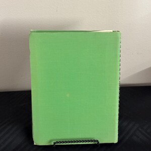May include: A green hardcover book with a white spine. The book is standing upright on a black surface.
