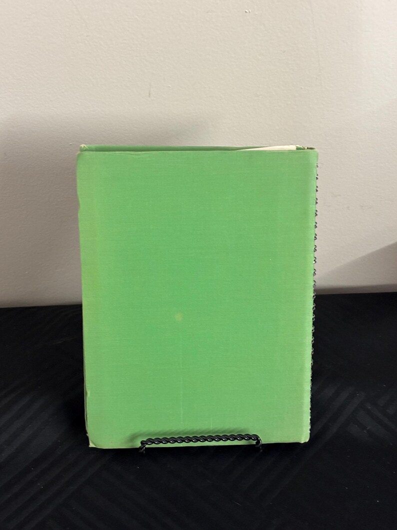 May include: A green hardcover book with a white spine. The book is standing upright on a black surface.