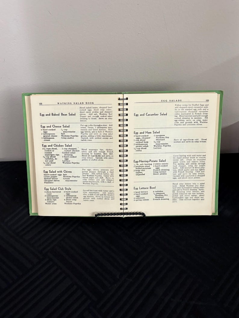 May include: A vintage Watkins Salad Book with recipes for egg salads. The book is open to a page with recipes for Egg and Baked Bean Salad, Egg and Cheese Salad, Egg and Chicken Salad, Egg Salad with Chives, Egg Salad Club Style, Egg and Cucumber Salad, Egg and Ham Salad, Egg-Herring-Potato Salad, and Egg Lettuce Bowl.
