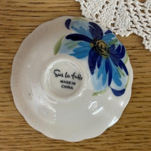 Puede incluir: Un peque&ntilde;o plato de cer&aacute;mica blanco con un dise&ntilde;o floral azul. El plato tiene un borde festoneado y el texto "Sur la table MADE IN CHINA" est&aacute; impreso en la parte inferior. La flor tiene p&eacute;talos azules y hojas verdes.
