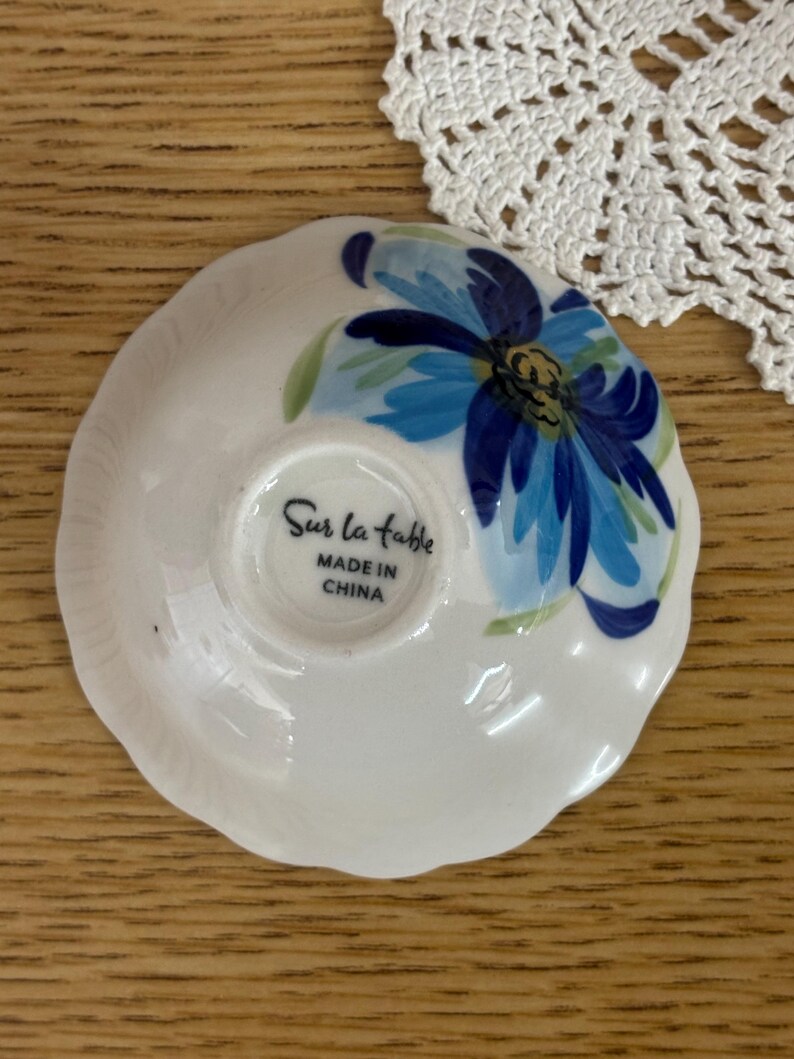 Puede incluir: Un peque&ntilde;o plato de cer&aacute;mica blanco con un dise&ntilde;o floral azul. El plato tiene un borde festoneado y el texto "Sur la table MADE IN CHINA" est&aacute; impreso en la parte inferior. La flor tiene p&eacute;talos azules y hojas verdes.