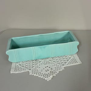 May include: A rectangular, light turquoise ceramic planter with a smooth, glossy finish. The exterior features vertical indented lines along the sides and a scalloped edge design on the short ends, creating a clean, structured appearance.