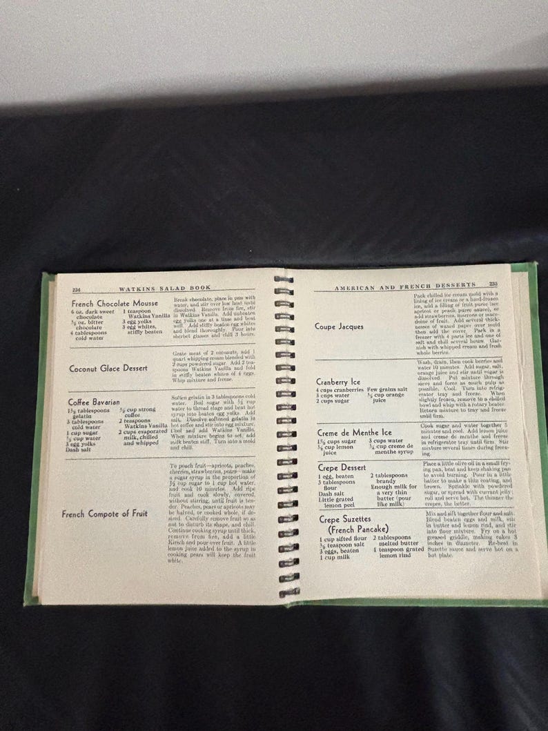 May include: A vintage Watkins Salad Book cookbook open to pages with recipes for French Chocolate Mousse, Coconut Glace Dessert, Coffee Bavarian, French Compote of Fruit, Coupe Jacques, Cranberry Ice, Creme de Menthe Ice, Crepe Dessert, and Crepe Suzettes (French Pancake).