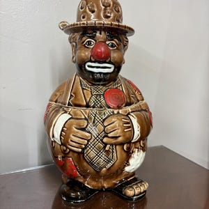 May include: A brown ceramic clown cookie jar with a red nose, a brown hat, and a checkered tie. The jar is in the shape of a seated clown wearing a suit with red patches. The jar has a lid.