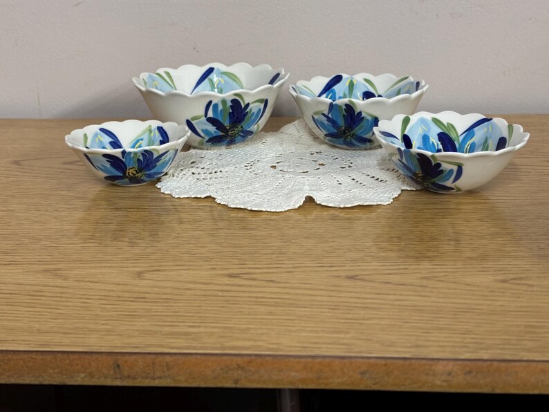 Puede incluir: Conjunto de cuatro cuencos de cer&aacute;mica blanca con bordes festoneados. Cada cuenco presenta un dise&ntilde;o floral azul pintado a mano con detalles verdes. Los cuencos est&aacute;n colocados sobre un tapete de encaje blanco.