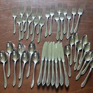 May include: A collection of silver-colored flatware, including forks, spoons, and knives, arranged on a wooden surface. The silverware has a classic design, with curved handles and a polished finish, suitable for formal dining.