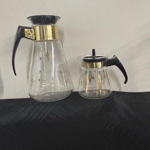 May include: A set of two glass coffee carafes with black and gold accents. The larger carafe has a starburst design and the smaller carafe has a geometric design.