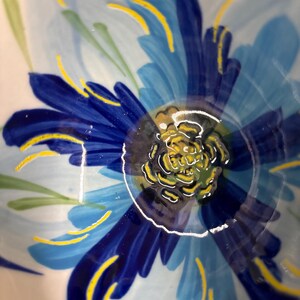 Puede incluir: Primer plano de un cuenco de cer&aacute;mica con un dise&ntilde;o floral pintado a mano. La flor presenta tonos de p&eacute;talos azules con detalles amarillos y un centro amarillo. Tambi&eacute;n hay hojas verdes pintadas en la superficie del cuenco. El cuenco tiene un acabado brillante.
