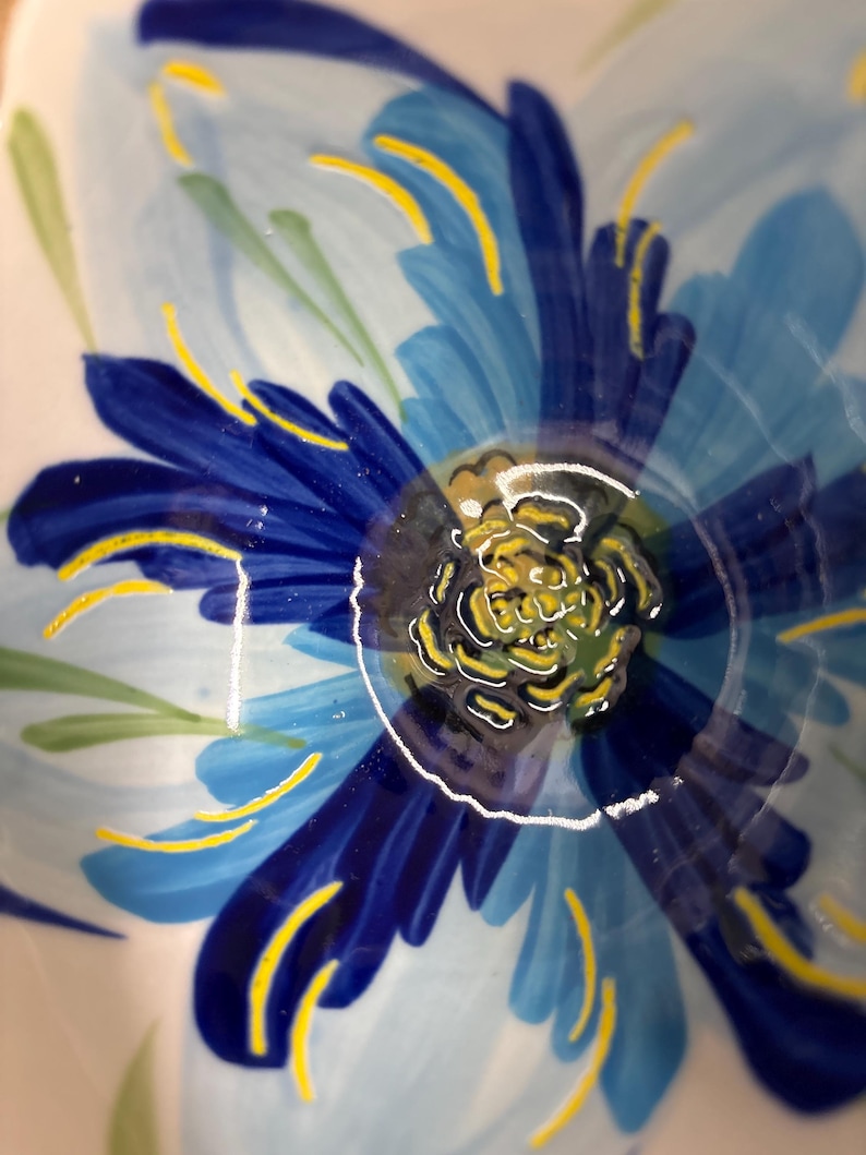 Puede incluir: Primer plano de un cuenco de cer&aacute;mica con un dise&ntilde;o floral pintado a mano. La flor presenta tonos de p&eacute;talos azules con detalles amarillos y un centro amarillo. Tambi&eacute;n hay hojas verdes pintadas en la superficie del cuenco. El cuenco tiene un acabado brillante.