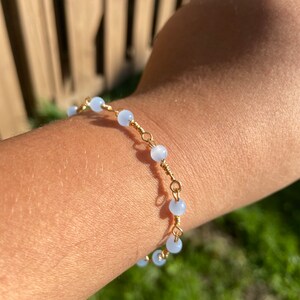 May include: A delicate bracelet featuring light blue beads and gold wire. The bracelet is worn on a person's wrist.