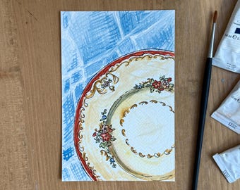 May include: Watercolor painting of a decorative plate with a red and gold rim and floral accents. The plate is set against a blue and white background. A paintbrush and paint tubes are visible.