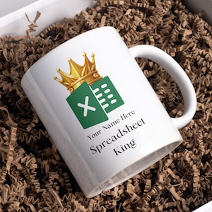 May include: White ceramic mug with a gold crown and a green Excel spreadsheet icon. The mug has the text "Your Name Here Spreadsheet King" printed on it. The mug is inside a white box filled with brown crinkle paper.