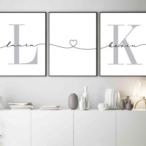 May include: Three black framed prints with white backgrounds. The left print features a large grey letter L with the word "laura" written in cursive above it. The middle print features a heart shape with a line extending to the left and right. The right print features a large grey letter K with the word "kevin" written in cursive above it.