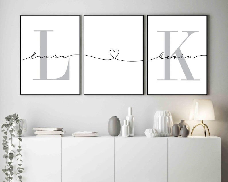 May include: Three black framed prints with white backgrounds. The left print features a large grey letter L with the word "laura" written in cursive above it. The middle print features a heart shape with a line extending to the left and right. The right print features a large grey letter K with the word "kevin" written in cursive above it.