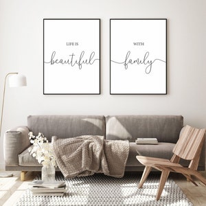 Op de afbeelding: Twee zwarte ingelijste prints met witte achtergronden en zwarte kalligrafie. De linker print zegt "Het leven is mooi" en de rechter print zegt "met familie". De prints hangen aan een witte muur boven een grijze bank met een bruine deken.