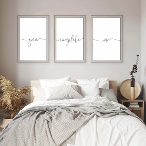 May include: Three white framed prints with black calligraphy on a white background. The prints read "you", "complete", and "me". The prints are hanging on a white wall above a white bed with a gray blanket.