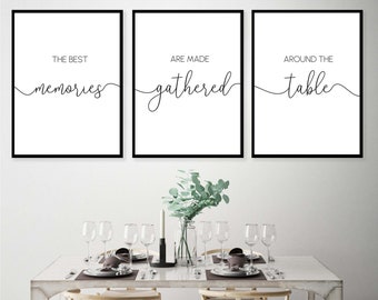 May include: Three black framed prints with white backgrounds and black calligraphy. The prints read "The best memories are made gathered around the table". The prints are hanging on a white wall above a white dining table with six chairs. The table is set with plates, cutlery, glasses, and a vase of green eucalyptus.