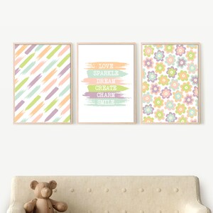 May include: Three framed prints with pastel colors. The left print has a pattern of diagonal stripes in shades of pink, green, and purple. The middle print has the words "LOVE SPARKLE DREAM CREATE CHARM SMILE" in white text on a background of pastel colored brushstrokes. The right print has a pattern of pastel colored flowers on a white background.