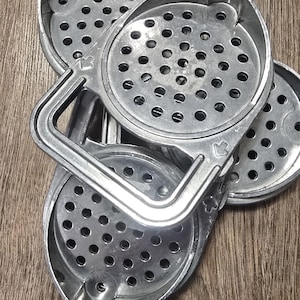 Puede incluir: Tres boles ralladores de metal de color plateado con un asa. Los boles son circulares con un fondo perforado y un borde elevado. El asa está unida al lado de los boles y tiene una forma curva.