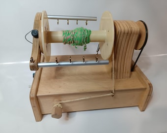 May include: A wooden yarn winder with a green and pink yarn skein on the spindle. The winder has a crank on the side and a small wooden handle.