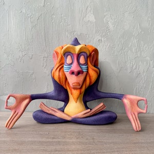 Puede incluir: Una figurita de mono de color morado, amarillo y naranja, sentada en una postura meditativa con los ojos cerrados. El mono tiene una larga melena naranja y una cara azul.