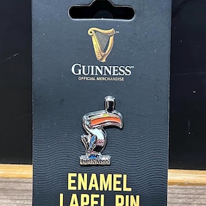 May include: A Guinness enamel lapel pin featuring a colorful toucan design. The pin is attached to a black card with the Guinness logo and the words "OFFICIAL MERCHANDISE". The card also reads "ENAMEL LAPEL PIN".