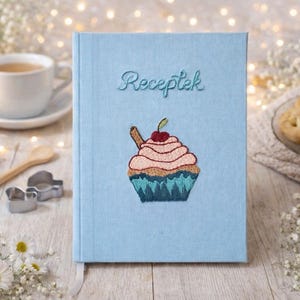Puede incluir: Un libro de recetas azul claro con la palabra "Receptek" bordada en la portada. Un diseño de cupcake con glaseado rosa, una cereza y una ramita de canela está bordado debajo del texto. El libro está rodeado de una taza de té, galletas y flores.