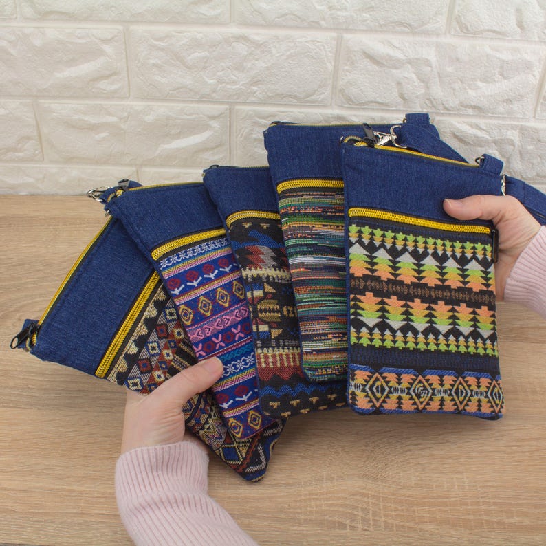 A person holding several handmade crossbody bags with colorful, ethnic-style tapestry pockets and golden color zippers, displayed against a wooden surface and white textured wall.