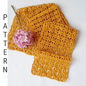 May include: A crocheted scarf in a mustard yellow color. The scarf is made with a delicate open stitch pattern and is laid out on a white surface.