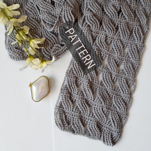 May include: A gray crocheted scarf with a lacy pattern. The scarf is laid out on a white surface with a small decorative box and a sprig of white flowers.
