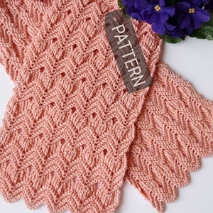 May include: A pink crocheted scarf with a delicate, lacy pattern. The scarf is folded in half and has a scalloped edge. The word "PATTERN" is written on a brown tag.